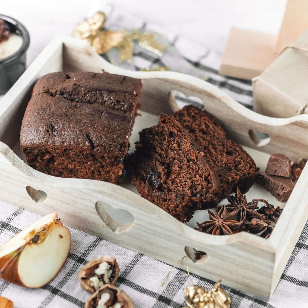Ein Holzkästchen gefüllt mit einem Stück Schokoladenkuchen und Schokoladenstückchen. Der Kuchen steht auf einem Tisch und um das Kästchen herum sind verschiedene Früchte wie Äpfel und Orangen angeordnet. Das Bild zeigt eine Nahaufnahme des Schokoladenkuchens, mit einem verlockenden Aussehen. Ein inspirierendes Bild für ein köstliches Schokokuchen Rezept